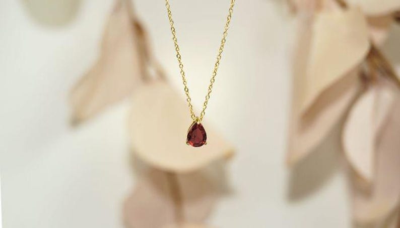 May include: A gold-colored necklace with a delicate chain and a teardrop-shaped, deep red gemstone pendant. The pendant is the focal point, suspended gracefully against a soft, blurred background. Jewelry.
