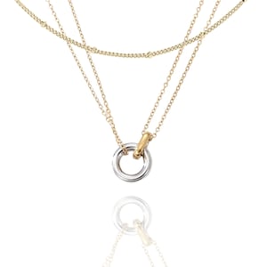 May include: A layered necklace with gold and silver tones. The necklace features three chains, with a silver and gold pendant. The pendant is a double ring design, with one silver ring and one gold ring.