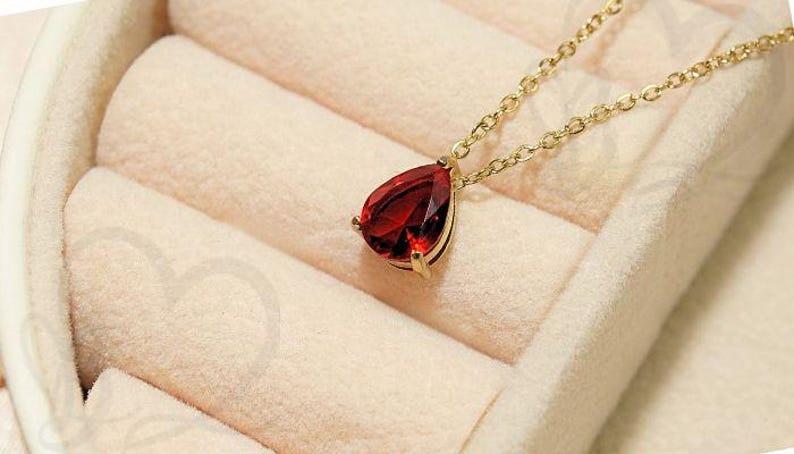 May include: A gold-colored necklace with a teardrop-shaped, deep red gemstone pendant. The pendant is set in a gold-colored bezel and hangs from a delicate chain. The necklace rests on a soft, light-colored fabric.