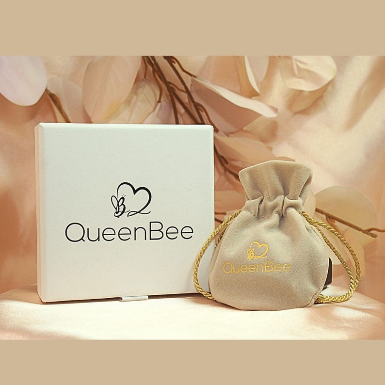 May include: A white box and a beige drawstring pouch with the "QueenBee" logo. The box has a black heart and bee logo with the words "QueenBee" below. The pouch has a gold drawstring and the "QueenBee" logo.