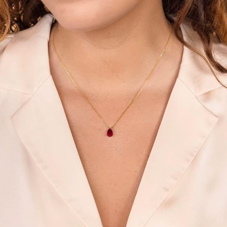 May include: A delicate gold necklace featuring a small, teardrop-shaped, deep red gemstone pendant. The necklace is displayed against a light-colored background, highlighting the pendant's color and the chain's subtle shine. The chain is thin and elegant.