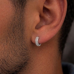 May include: A small silver-toned hoop earring featuring a row of clear, sparkling stones embedded along the front surface. The circular jewellery piece is secured through the earlobe, showcasing a polished metallic finish and a series of round gems.