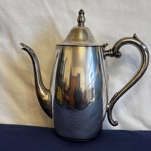 May include: A polished silver coffee pot with a curved spout and ornate handle. The pot has a lid with a decorative finial. The reflective surface mirrors the surrounding environment. The coffee pot is likely vintage.