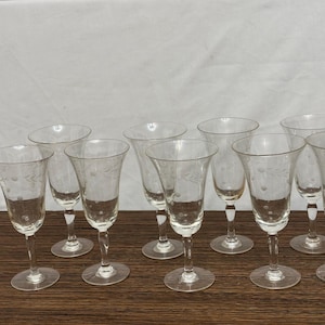 May include: Set of ten clear glass wine glasses with etched floral designs. The glasses are arranged in two rows on a wooden surface.
