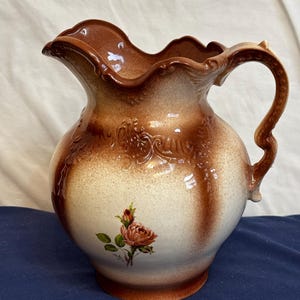 Könnte beinhalten: Eine Keramikkrug in Braun- und Cremetönen. Er hat ein Blumenmuster und einen dekorativen Griff. Der Rand ist gewellt. Der Krug ist etwa 25 cm hoch.