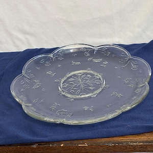 May include: Clear glass serving plate with a scalloped edge and floral embossed detailing. The center features a circular design with a starburst pattern. The plate is designed for serving food and is a decorative piece.