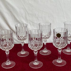 May include: A set of six clear crystal wine glasses, each featuring a diamond-cut design. The glasses have a stemmed base and a circular foot. One glass has a gold sticker. They are displayed on a red surface.