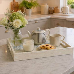 Puede incluir: Una bandeja rectangular con un diseño de olas azules y blancas contiene una tetera, una taza, una vela y galletas. Un jarrón de rosas blancas y vegetación se encuentra en la bandeja. La tetera tiene un diseño a rayas blancas y azules.