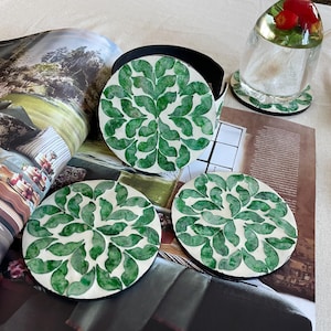 Pode incluir: Um conjunto de quatro bases redondas com fundo branco e design de folhas verdes. As bases são exibidas com um suporte a condizer, um copo e uma revista.