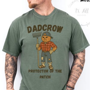 May include: Moss green t-shirt with a graphic of a pumpkin-headed scarecrow holding a rake. The text "DADCROW PROTECTOR OF THE PATCH" is above and below the illustration. The shirt is a comfortable fit.