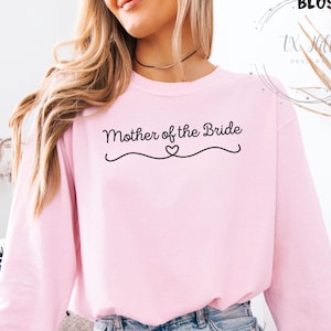 May include: A light pink sweatshirt with the text "Mother of the Bride" in black script. The text is above a decorative line with a heart in the middle. The sweatshirt has long sleeves and a crew neck.