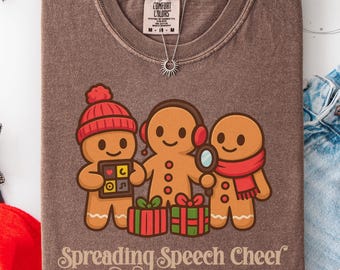 May include: Brown Comfort Colors t-shirt with a gingerbread family design. The gingerbread people wear a red hat, headphones, and scarf. The text "Spreading Speech Cheer" is printed on the shirt. A silver necklace with a sun pendant is visible.