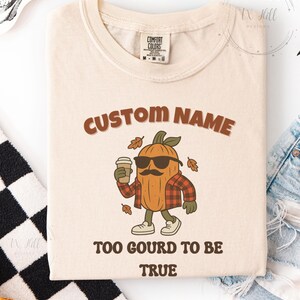 May include: Ivory-colored t-shirt with the text "CUSTOM NAME" above a cartoon pumpkin wearing sunglasses and a plaid shirt, holding a coffee cup. The text "TOO GOURD TO BE TRUE" is below the pumpkin. The shirt has a Comfort Colors tag.