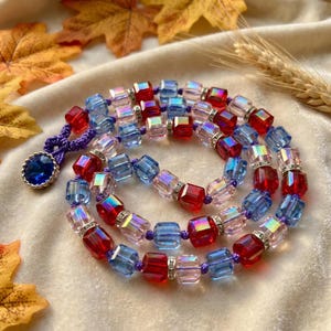May include: A necklace featuring alternating cube-shaped beads in red, light blue, and iridescent clear glass, separated by small silver-toned metal spacers with embedded crystals. The beads are strung on a purple cord, ending in a dark blue pendant.