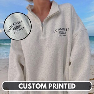 May include: A light gray sweatshirt with a quarter-zip collar and the words "Sunkissed Coconut" printed in black. The sweatshirt has a relaxed fit and a small embroidered design above the text. The bottom of the image has the words "CUSTOM PRINTED".