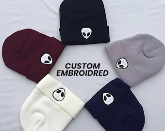 May include: Five beanies in different colors with an alien face embroidered on the front. The beanies are navy blue, burgundy, gray, white, and dark blue. The text "CUSTOM EMBROIDERED" is in the center of the image.