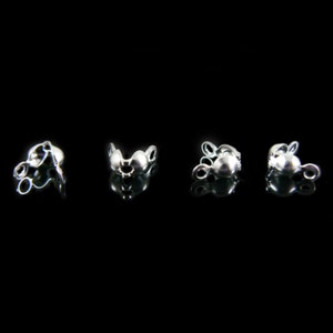 May include: Four small, silver-colored jewelry findings are arranged in a row against a black background. Each finding has a rounded shape with a small loop on each side, and a larger, central bead-like element.