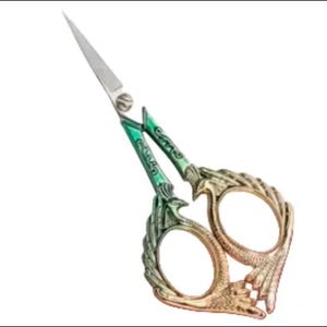May include: Decorative embroidery scissors with a silver blade and ornate handles. The handles feature a green and gold design with a bird or phoenix motif. The scissors are designed for detailed cutting and crafting.