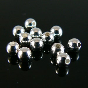 May include: A group of silver-colored metal beads, all the same size and shape, are scattered on a black surface. The beads are round and have a smooth, shiny finish.