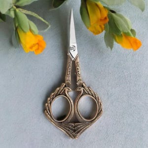 May include: Ornate bronze-colored scissors with a decorative bird design on the handles. The scissors have sharp, silver blades and are set against a soft blue background with yellow flowers.