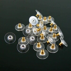 May include: A collection of clear, disc-shaped earring backs with gold-colored posts and silver-colored centers. The earring backs are scattered on a dark surface, showcasing their transparent design and metallic accents.