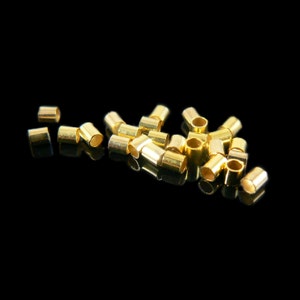 May include: A pile of gold-colored metal jump rings, scattered on a black surface. The rings are small and cylindrical, with a smooth finish.