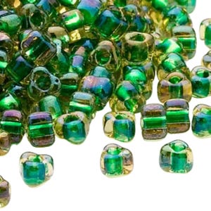 Puede incluir: Primer plano de un montón de cuentas de vidrio verde iridiscente. Las cuentas son cilíndricas y tienen una mezcla de tonos verdes, dorados y azules. Las cuentas están dispersas, algunas enfocadas y otras borrosas.