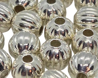 May include: A collection of round, metallic silver beads featuring a vertical ribbed or fluted texture. Each bead has a central hole for stringing. The surface of the beads is highly reflective, creating bright highlights and deep shadows.