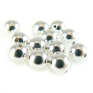 May include: A group of silver metal beads, each with a small hole in the center. The beads are round and shiny, and they are scattered on a white surface.