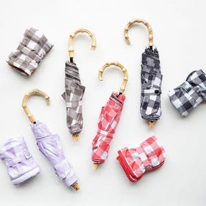 Puede incluir: Un conjunto de ocho mini paraguas con un patrón a cuadros en varios colores, incluyendo rojo, negro, blanco y morado. Los paraguas tienen mangos de bambú y están plegados.