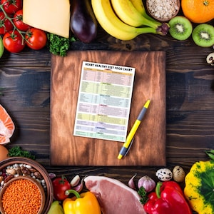 May include: A heart-healthy diet food chart with a yellow pen on a wooden cutting board surrounded by various fruits, vegetables, and other foods. The chart lists different food groups and their nutritional value.