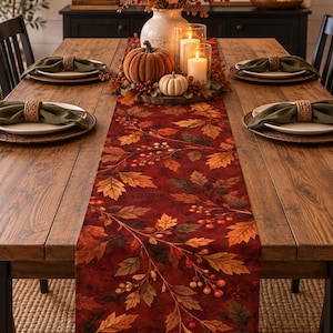 May include: A long rectangular table runner featuring a deep red background with a printed pattern of autumn leaves and small berries. The fabric runner is centered on a wooden dining table, decorated with pumpkins and candles in glass holders.