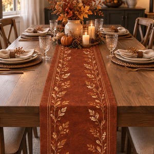 May include: A rectangular table runner in a deep rust-orange color featuring a repeating vertical pattern of light-colored leaves and berries along both sides, centered on a wooden dining table set with plates, napkins, and candles.