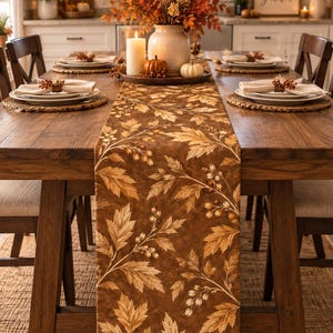 May include: A long rectangular table runner featuring a brown fabric base with a repeating pattern of light brown and tan autumn leaves and small berries. The runner is draped down the center of a wooden dining table, showcasing the botanical print.
