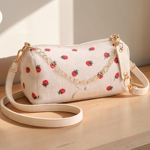 Könnte beinhalten: Cremefarbene Cord-Umhängetasche mit einem Muster aus roten Erdbeeren. Die Tasche hat einen goldfarbenen Kettenriemen und einen cremefarbenen, verstellbaren Schulterriemen. Die Tasche hat eine rechteckige Form mit abgerundeten Kanten.