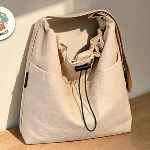 Puede incluir: Un bolso de hombro beige con cierre de cordón y un bolsillo frontal grande. El bolso tiene una solapa que se pliega sobre la parte superior. El bolso está hecho de un material suave y texturizado. Un logotipo circular con el texto "CARRY FREELY" es visible.