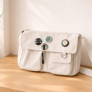 Pode incluir: Bolsa mensageiro de lona creme com fecho de aba, dois bolsos frontais e uma alça de ombro ajustável. A bolsa é decorada com vários pinos decorativos. A bolsa está sobre uma superfície de madeira.