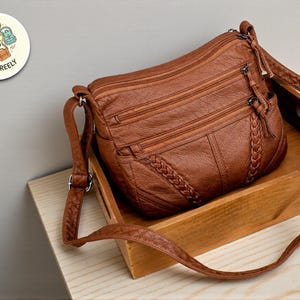 Puede incluir: Un bolso bandolera marrón con múltiples compartimentos con cremallera y un detalle trenzado. El bolso tiene una correa ajustable y está hecho de un material texturizado. El bolso está sobre una bandeja de madera.