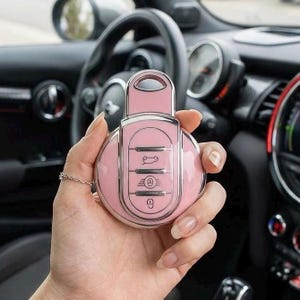 May include: A pink car key fob with silver detailing, held in a hand. The key has buttons for locking, unlocking, and other functions. The background shows a car's interior, including the steering wheel and dashboard.