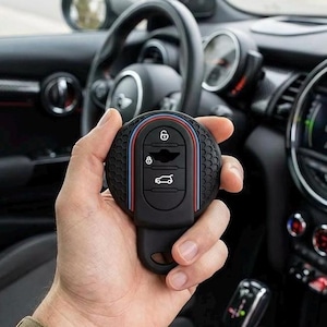 May include: Black car key fob cover with red, white, and blue stripes. The key fob has a textured surface and features the car's logo and key functions. The background shows the car's interior, including the steering wheel and dashboard.