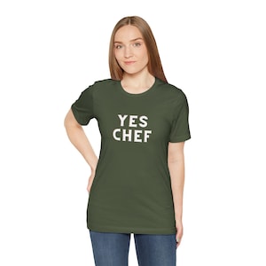 May include: An olive green t-shirt with the text "YES CHEF" in white block letters. The shirt is a crew neck style and is being worn by a person. The person is wearing blue jeans.