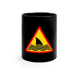 May include: Black ceramic mug with a yellow triangle warning sign with a black shark fin and waves inside. The sign has a red border.