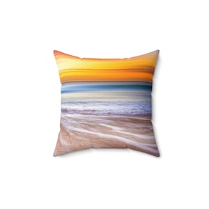 May include: A decorative pillow with a sunset over the ocean scene. The pillow features a gradient of orange, yellow, and blue colors, with white waves crashing on the sandy shore.