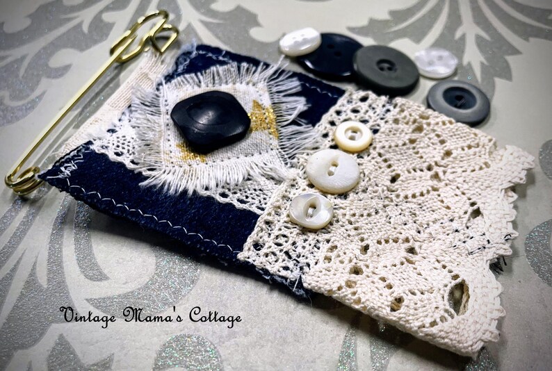 Puede incluir: Una pieza textil artesanal con un dise&ntilde;o en azul marino y encaje crema, adornada con un bot&oacute;n negro grande y varios botones blancos m&aacute;s peque&ntilde;os. Tambi&eacute;n hay un imperdible dorado y varios botones. El texto "Vintage Mama's Cottage" es visible.