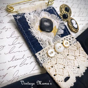 Puede incluir: Broche textil artesanal con base de mezclilla azul marino, adornado con un bot&oacute;n negro, encaje blanco y peque&ntilde;os botones blancos. Un imperdible dorado y pendientes vintage con detalles en blanco y negro completan el dise&ntilde;o. El texto "Vintage Mama's Cottage" es visible.