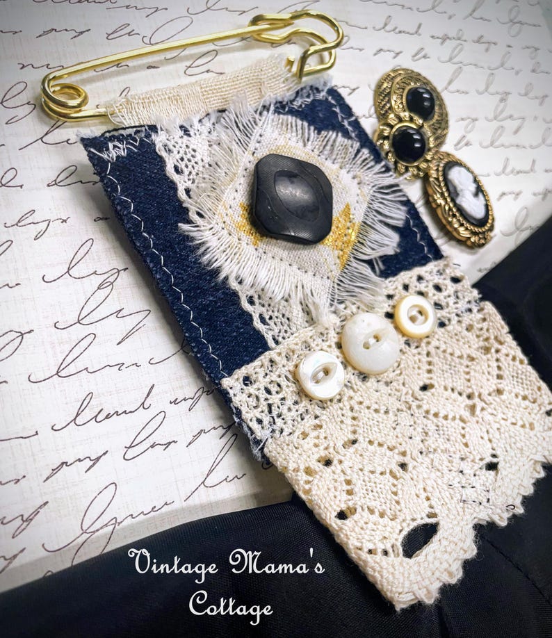Puede incluir: Broche textil artesanal con base de mezclilla azul marino, adornado con un bot&oacute;n negro, encaje blanco y peque&ntilde;os botones blancos. Un imperdible dorado y pendientes vintage con detalles en blanco y negro completan el dise&ntilde;o. El texto "Vintage Mama's Cottage" es visible.
