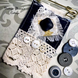 Puede incluir: Una pieza textil artesanal con fondo azul marino, adornada con encaje blanco, botones y un bot&oacute;n negro grande. Un imperdible dorado asegura la parte superior. Varios botones adicionales en varios tama&ntilde;os y tonos de gris y blanco est&aacute;n dispersos cerca.