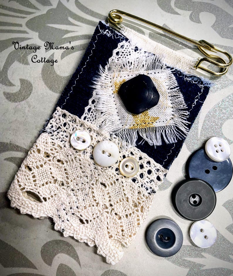Puede incluir: Una pieza textil artesanal con fondo azul marino, adornada con encaje blanco, botones y un bot&oacute;n negro grande. Un imperdible dorado asegura la parte superior. Varios botones adicionales en varios tama&ntilde;os y tonos de gris y blanco est&aacute;n dispersos cerca.