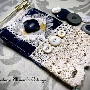 Puede incluir: Un alfiler textil hecho a mano con una base azul marino, adornado con un bot&oacute;n negro, encaje y un imperdible dorado. Varios botones vintage de varios tama&ntilde;os y colores est&aacute;n dispersos cerca. El texto "Vintage Mama's Cottage" es visible.