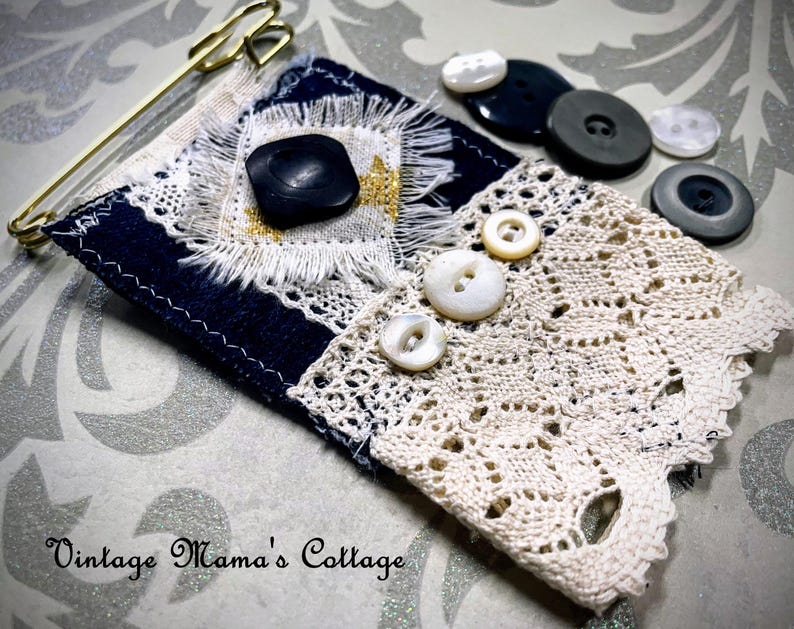 Puede incluir: Un alfiler textil hecho a mano con una base azul marino, adornado con un bot&oacute;n negro, encaje y un imperdible dorado. Varios botones vintage de varios tama&ntilde;os y colores est&aacute;n dispersos cerca. El texto "Vintage Mama's Cottage" es visible.